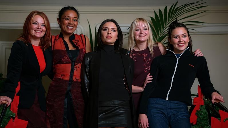 Cinco mulheres jovens estão alinhadas em escada interna decorada com guirlandas verdes e laços vermelhos. Elas vestem roupas variadas, incluindo vestido preto, saia de couro, blusa vermelha, e jaqueta preta. Ao fundo, parede branca e planta verde alta.
