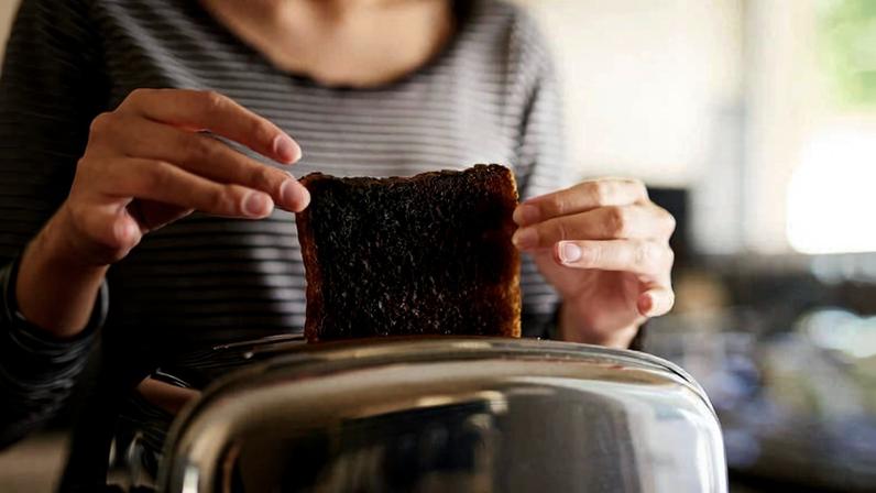Pessoa segurando uma fatia de pão visivelmente queimada ao retirá-la de uma torradeira metálica em ambiente doméstico.