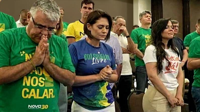 Dez pessoas em pé, algumas com as mãos unidas em oração, vestindo camisetas verdes, azuis e brancas com mensagens. Ambiente interno com cadeiras e parede ao fundo.