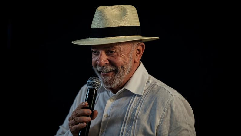 Homem idoso com barba branca usa chapéu branco com faixa preta e camisa social branca enquanto segura microfone. Fundo desfocado mostra várias pessoas e câmeras, sugerindo evento com público.