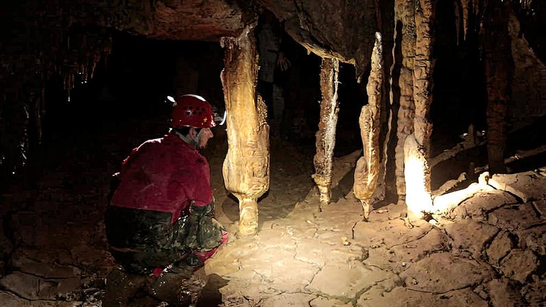 Pessoa vestindo roupa vermelha e capacete com lanterna agachada dentro de caverna, iluminando estalactites e estalagmites no chão de pedra rachada.