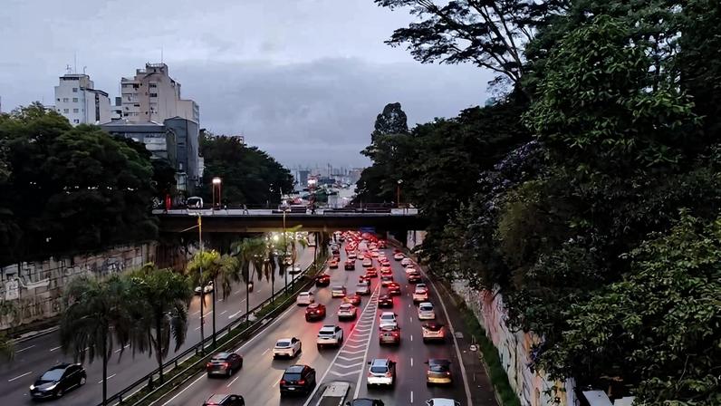 Veículos congestionados na Marginal Pinheiros em São Paulo sob céu nublado. Faixa da direita está mais lenta, com carros parados e lentos, enquanto faixa da esquerda tem fluxo mais livre. Vegetação densa e prédios altos cercam a via.