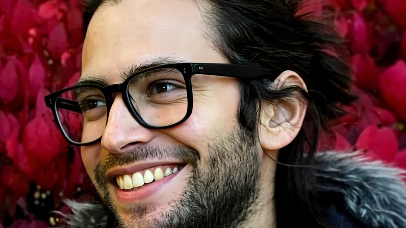 Homem jovem com barba rala e cabelos presos usa óculos de armação escura. Ele sorri olhando para o lado, com fundo de folhas vermelhas vibrantes.