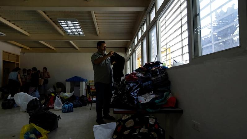 Homem de máscara organiza pilha de roupas sobre mesa próxima a janelas grandes em sala ampla. No fundo, grupo de pessoas conversa próximo a sacolas e pacotes no chão.