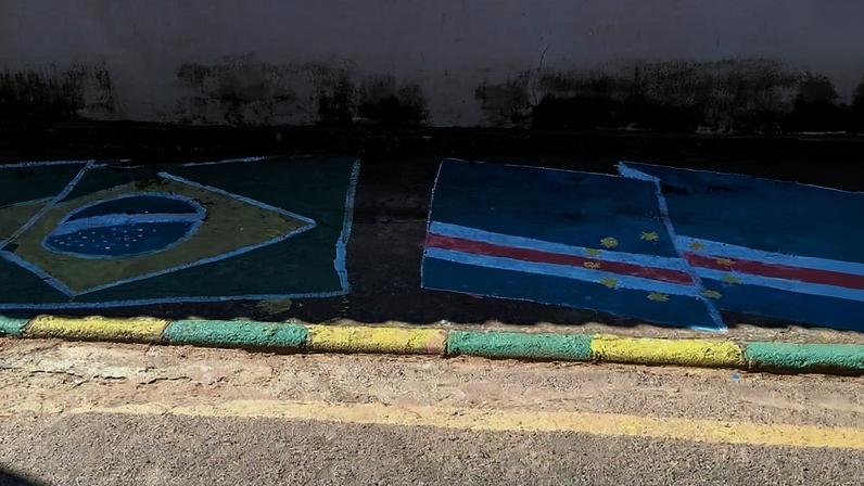 Bandeira do Brasil pintada no chão à esquerda, com as cores verde, amarelo, azul e branco. À direita, bandeira de Pernambuco pintada no chão, com fundo azul, faixa branca e vermelha, e estrelas amarelas. Calçada com meio-fio pintado em amarelo e verde, parede branca ao fundo com manchas de sujeira.