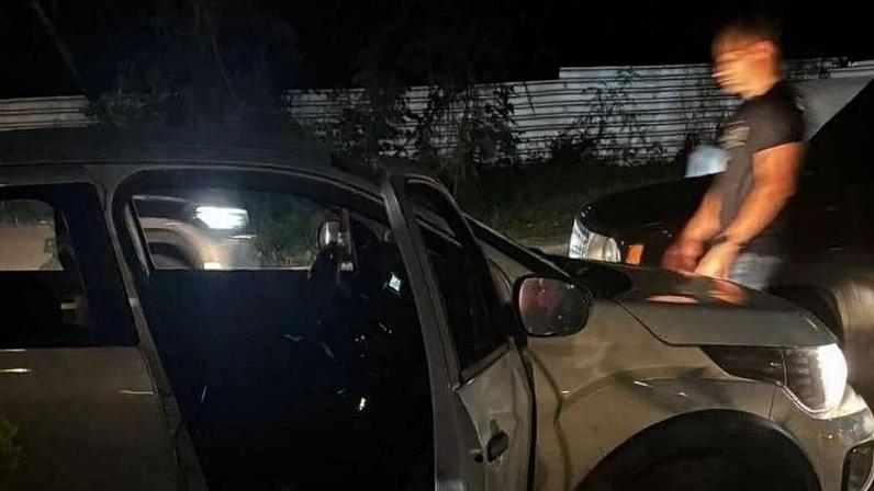 Veículo prata estacionado à noite com a porta dianteira esquerda aberta. Duas pessoas estão próximas ao carro, uma à esquerda e outra à direita, ambas em pé. Ambiente externo com iluminação baixa e vegetação ao fundo.