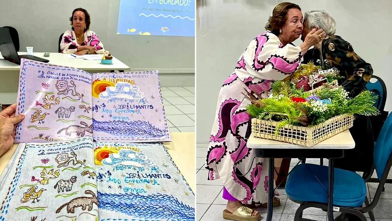 Duas mulheres interagem em sala de aula; uma delas está sentada atrás de uma mesa com um laptop e bordados coloridos à frente, enquanto a outra se inclina para cumprimentá-la com um beijo no rosto, ao lado de uma mesa com arranjos florais em cesto.