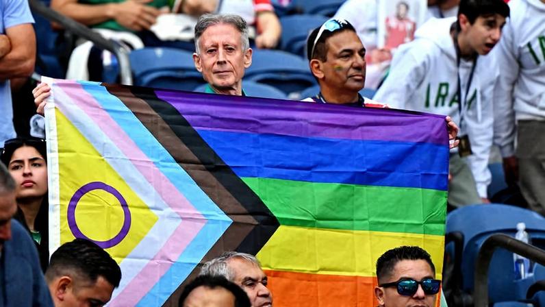 Duas pessoas seguram uma bandeira do orgulho LGBTQIA+ com as cores do arco-íris e faixas adicionais representando diversidade e inclusão. Elas estão em meio a uma multidão sentada em arquibancadas, algumas pessoas vestem roupas com a palavra 'Irã'.