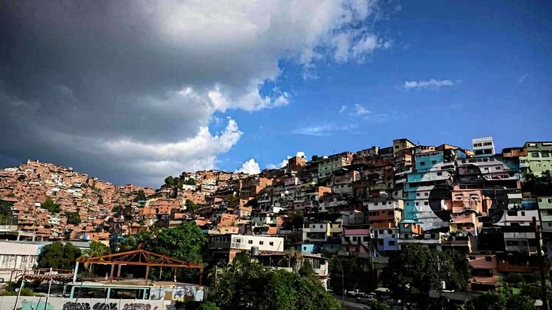 Vista panorâmica de um morro densamente ocupado por casas coloridas de diferentes tamanhos. O céu apresenta nuvens escuras à esquerda e céu azul com poucas nuvens à direita. Vegetação e algumas construções estão na base do morro.
