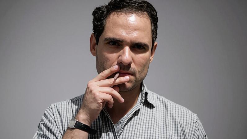 Homem de meia-idade com cabelo curto e escuro, vestindo camisa xadrez de mangas compridas arregaçadas, fuma um cigarro com a mão direita próxima à boca. A mão esquerda está no bolso da calça escura. Fundo branco liso.