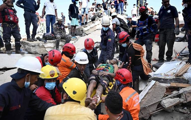 Equipe de resgate com capacetes coloridos e coletes salva vítima deitada em maca entre escombros de concreto. Pessoas observam ao fundo em área destruída sob céu claro.