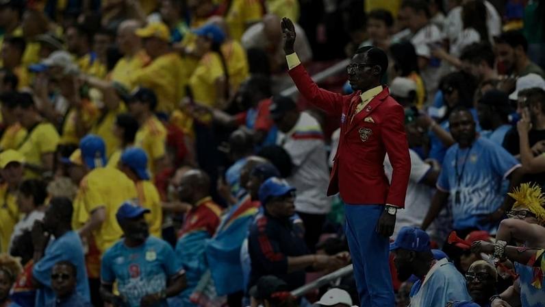 Homem vestido com paletó vermelho, calça azul e gravata amarela levanta o braço em estádio lotado. Público ao redor usa camisetas amarelas e azuis, muitos com bonés e acessórios coloridos.