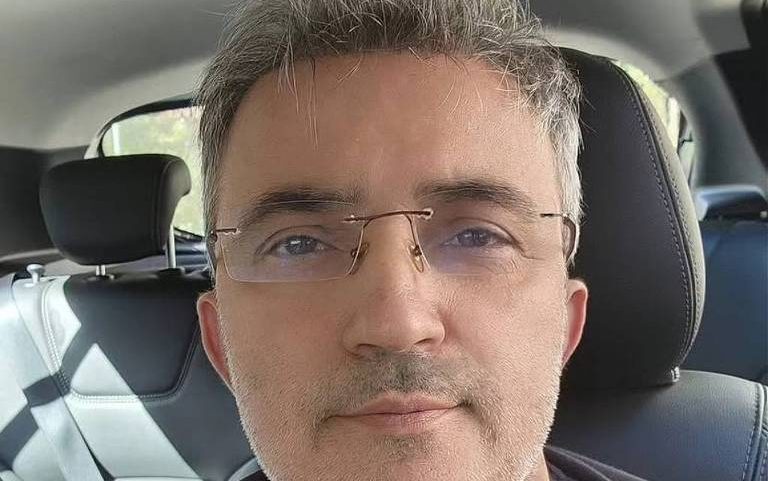 Homem de cabelos grisalhos e óculos finos está sentado no banco dianteiro de um carro. Ele veste camiseta preta com estampa de boca aberta mostrando dentes e lábios pintados com as cores do arco-íris.