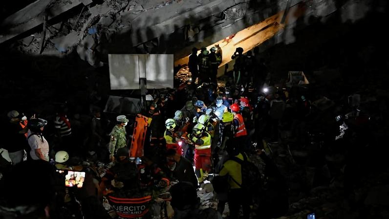 Equipe de resgate com capacetes e coletes refletivos trabalha em escombros à noite, iluminada por lanternas e holofotes. Pessoas ao redor registram a operação com celulares.