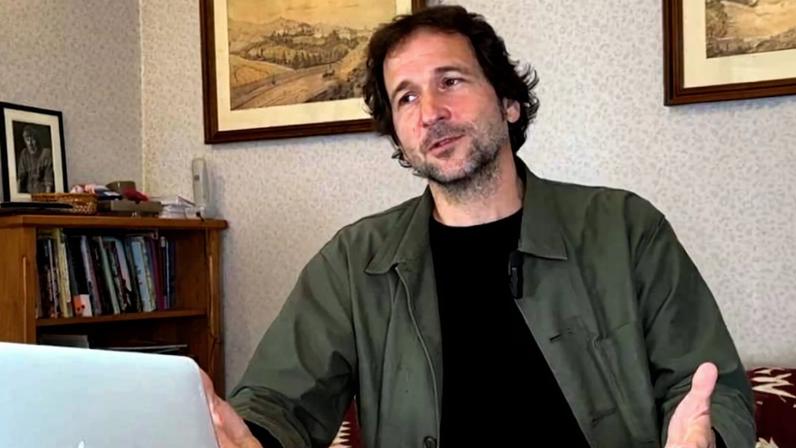 Homem de cabelos castanhos e barba rala sentado à mesa de madeira com laptop Apple aberto à sua frente. Ele veste jaqueta verde e camiseta preta, gesticulando com as mãos. Ao fundo, estante com livros, abajur e quadros nas paredes.