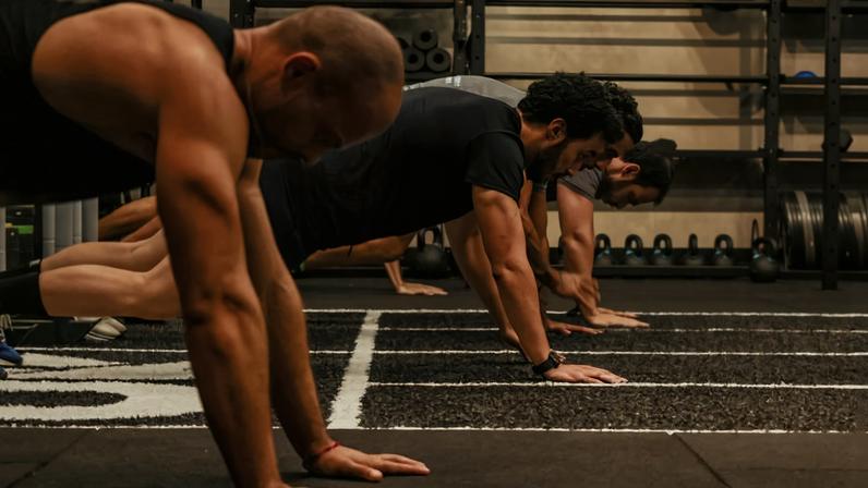 Três homens alinhados fazem flexões apoiados com as mãos no chão de academia. Ao fundo, prateleiras com bolas medicinais, halteres e pesos.
