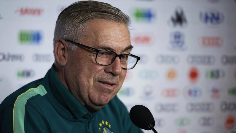 Homem de cabelos grisalhos e óculos escuros fala em microfone durante entrevista. Ele veste jaqueta verde da seleção brasileira com estrelas amarelas no peito. Fundo desfocado com logotipos.