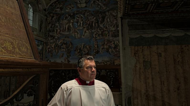 Grupo de 40 meninos e homens vestidos com túnicas brancas e saias roxas posam em fileiras diante do altar da Capela Sistina. Ao fundo, a grande pintura do Juízo Final cobre a parede, mostrando figuras humanas em várias posições e tons de azul e marrom.