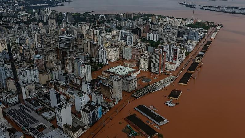 Vista aérea de Porto Alegre inundada, com prédios e ruas submersos em água barrenta. O centro da cidade e o cais do porto estão cobertos pela enchente, com apenas os telhados de algumas estruturas visíveis. O rio Guaíba, também marrom, se estende ao fundo, sob um céu nublado