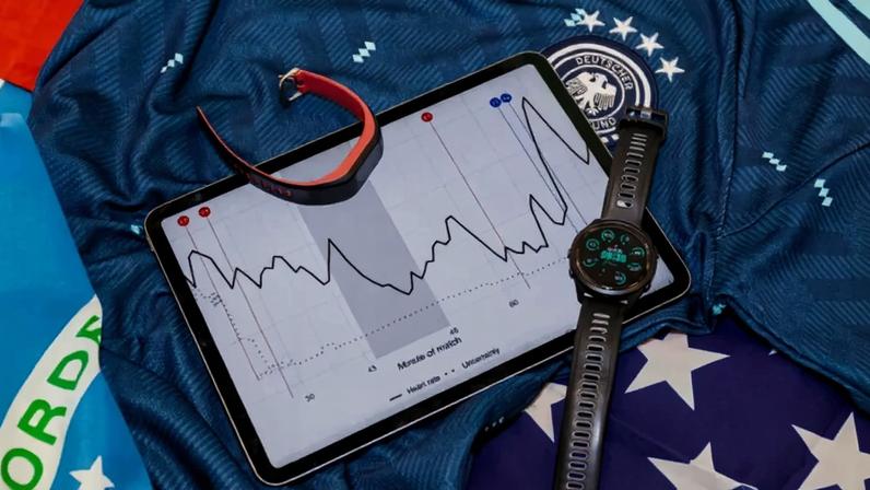 Tablet com gráfico de batimentos cardíacos durante uma partida, um relógio inteligente e uma pulseira fitness sobre uma camisa de futebol da Alemanha e bandeiras do Brasil e EUA