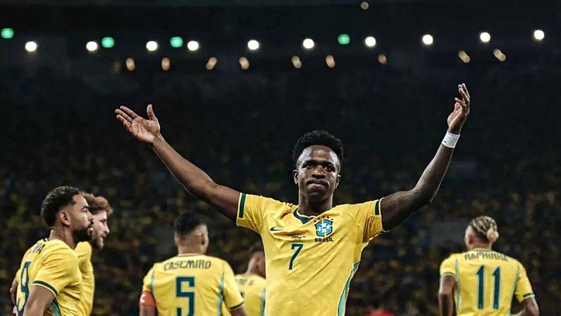 Vini Jr., com as mãos para o alto, acompanhado de outros jogadores da seleção brasileira de costas, todos uniformizados, em um estádio de futebol lotado