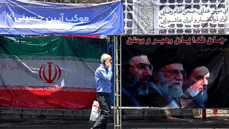 Um homem caminha ao lado da bandeira nacional do Irã e de uma faixa com as imagens do falecido fundador da Revolução Islâmica, o líder supremo Aiatolá Ruhollah Khomeini (à direita), do falecido líder supremo iraniano Ali Khamenei e de seu filho, o atual líder supremo Mojtaba Khamenei (à esquerda), erguidas ao longo de uma rua na Praça Vanak, em Teerã, em 10 de junho de 2026 - (crédito: ATTA KENARE / AFP)