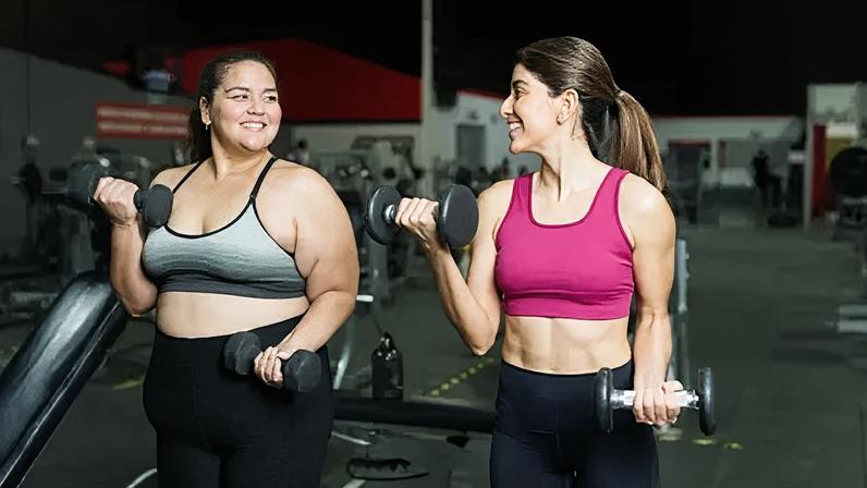 Getty Images Two women in gym gear stand next to each other and lift dumbell weights. They are smiling and enjoying the workout. One of them is overweight, the other is a healthy weight and has visibly toned arms and abs.
