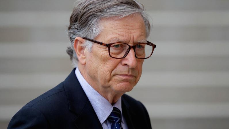 Bill Gates, de óculos e terno, sorri com cabelo grisalho levemente desarrumado pelo vento.
