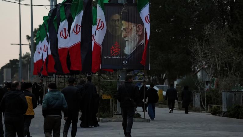 Pessoas caminham por uma rua com bandeiras iranianas e um cartaz com imagens do falecido Líder Supremo do Irã, o Aiatolá Ali Khamenei, em meio ao conflito entre os EUA e Israel com o Irã, em Teerã , Irã, 9 de março de 2026