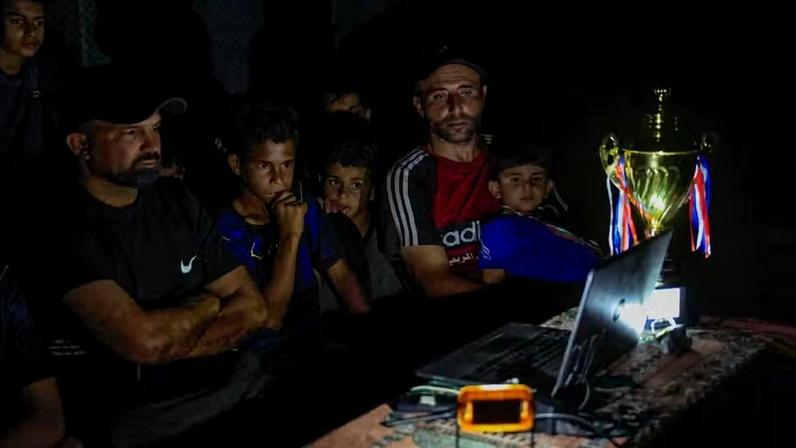 O ex-jogador de futebol palestino Fadi Al-Arawi assiste a uma partida da Copa do Mundo de 2026 transmitida em um laptop, junto com outras pessoas, em Khan Younis, no sul da Faixa de Gaza , em 13 de junho de 2026 — Foto: REUTERS/Haseeb Alwazeer