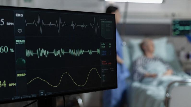 Monitor com indicadores de frequência cardíaca em primeiro plano, tendo ao fundo, um leito hospitalar com paciente deitado e um profissional de saúde de avental azul ao lado