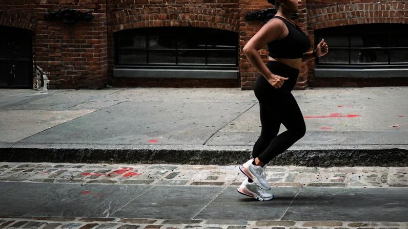 Mulher em atividade física corre por uma calçada, com edifício de tijolinhos ao fundo. Ela veste top e legging esportivos pretos e tênis brancos