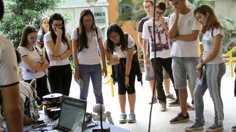 Vários estudantes em pé observam um robô pequeno no chão