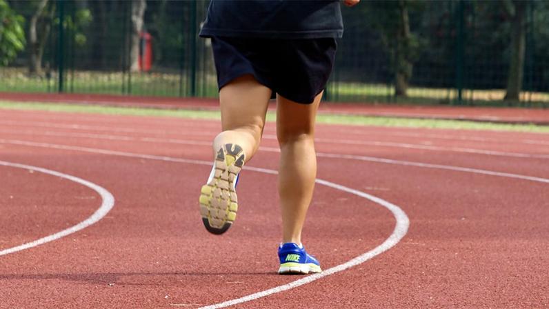 Mulher focalizada das pernas para baixo realiza corrida em pista de atletismo, usando camisa e calção preto, calçando um tênis azul