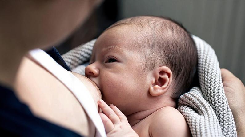 Mulher usando camisa azul amamenta bebê envolvido em um tecido de crochê que olha para o rosto de quem está dando de mamar a ele