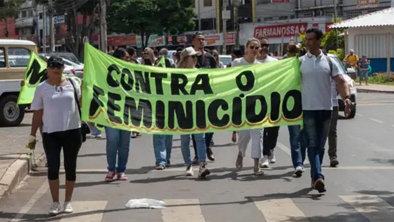 Pessoas carregam faixa em manifestação, em via pública, onde está escrito contra o feminicídio