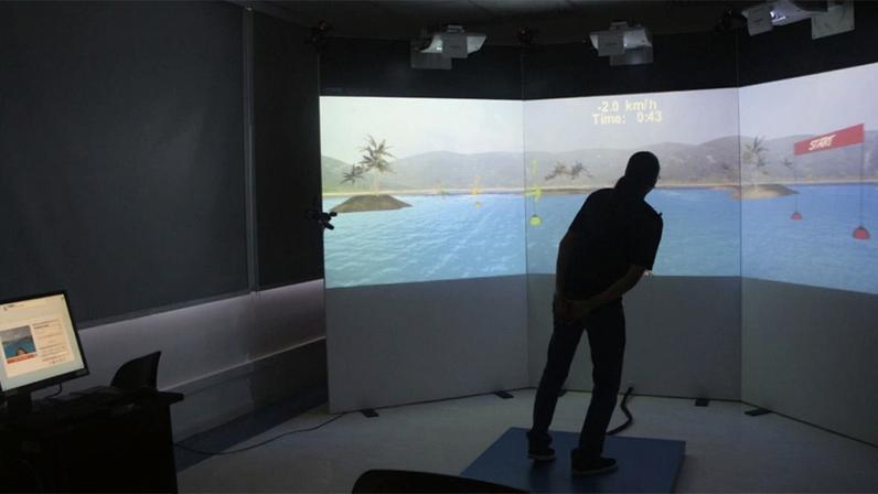 Pessoa sobre uma plataforma azul posicionada diante de um painel onde projetores instalados no teto projetam a tela de um jogo interativo, dentro de uma sala com cortinas fechadas e uma mesa com monitor de computador