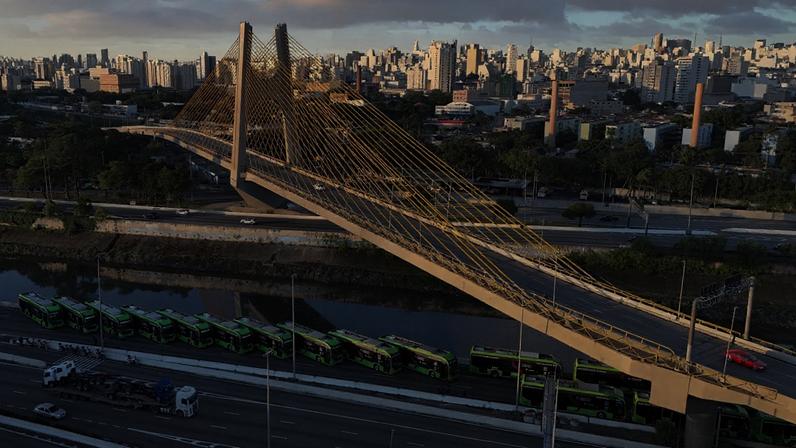 Ponte Estaiada Octávio Frias de Oliveira em São Paulo, com cabos amarelos e carros circulando. Abaixo, um rio e uma fila de ônibus verdes estacionados à beira da rodovia, com a cidade ao fundo.