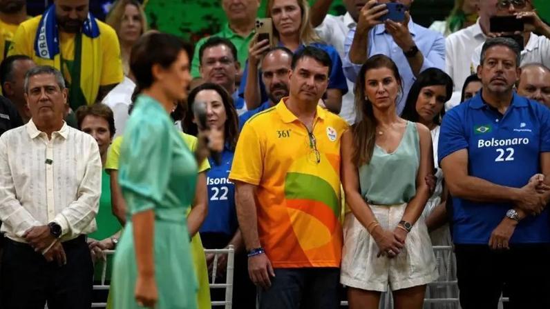 Michelle aparece em primeiro plano, desfocada, em vestido verde e falando ao microfone em um comício eleitoral de 2022. Ao fundo, Flávio e sua esposa observam Michelle ao lado de outras pessoas