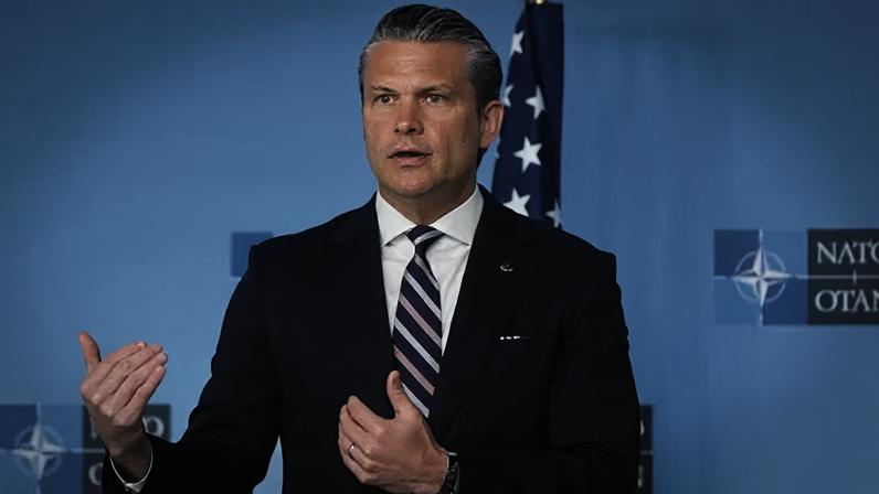 EPA Hegseth gestures with both of his hands as he stands in front of a Nato branded blue backdrop at a summit. He is wearing a dark blue suit, white shirt and white, blue and pink striped tie.