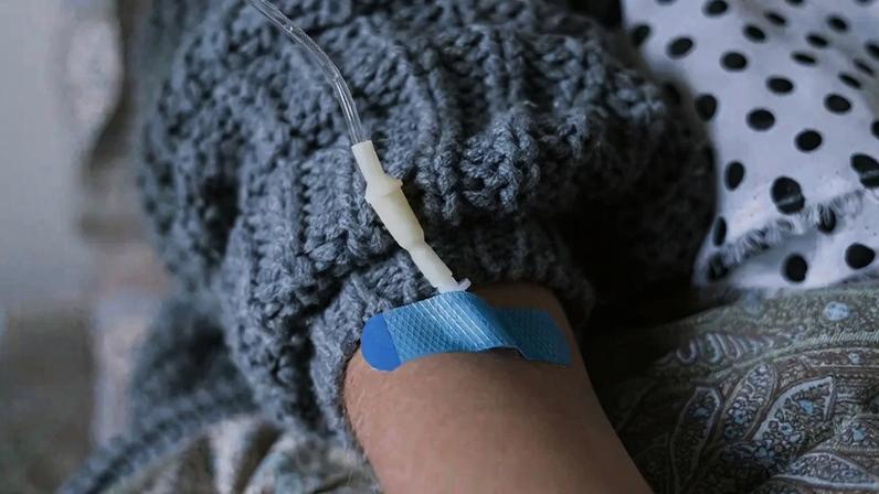 Braço de uma criança com um cateter intravenoso preso por um curativo azul, vestindo um suéter cinza e deitada em uma cama com lençóis estampados