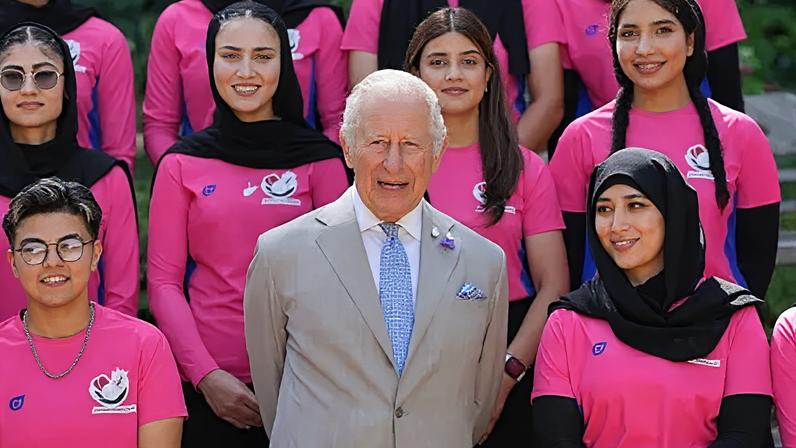 King Charles wearing a pale grey suit with white shirt and blue tie, with members of the Afghan women's cricket team - wearing hot pink T-shirts - on either side and behind him. All are looking ahead and smiling