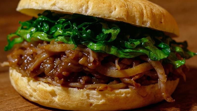 Em cima de uma mesa de madeira, está um pão de queijo grande recheado de pernil desfiado ao molho barbecue e tiras de rúcula.