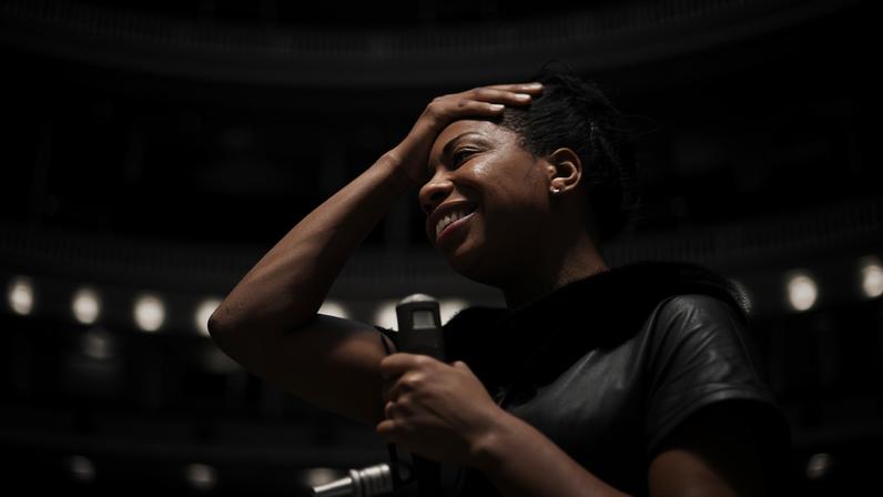 A photograph of a woman looking to the left, smiling. She has her right hand on her forehead and is holding a microphone in her right hand. In the background behind her you can see a large empty theater.
