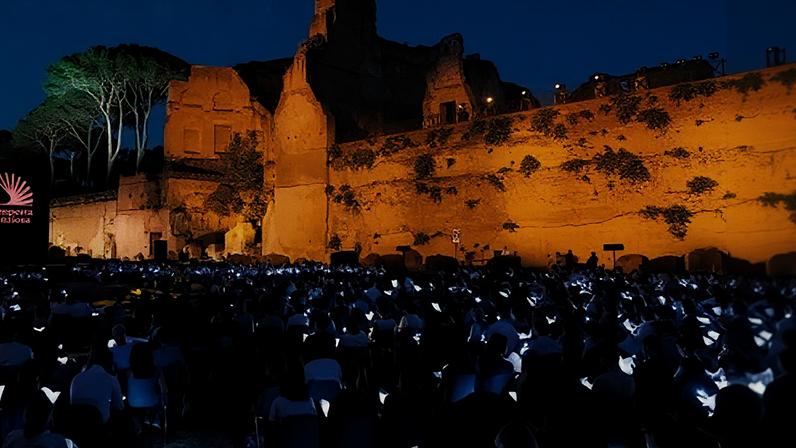 A large crowd sits in rows holding glowing devices at night before illuminated ancient ruins