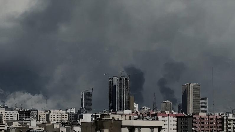 Fumaça escura sobe entre prédios de uma cidade, com montanhas ao fundo