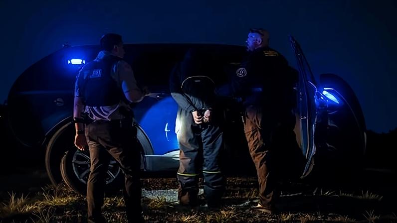 Two officers flanking a man who is handcuffed and facing a police vehicle. The sky is dark and a flashlight is illuminating the detainee’s hands.