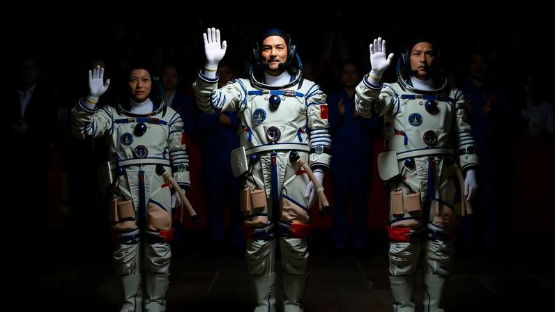 Três astronautas chineses, dois homens e uma mulher, em trajes espaciais brancos e azuis, acenam para uma multidão animada que segura bandeiras da China e de Hong Kong