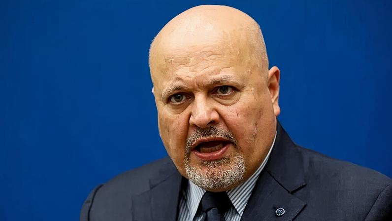 Reuters International Criminal Court Prosecutor Karim Khan speaks during an interview while sitting in front of a blue sign with blurry white writing and the symbol of the ICC - the scales of justice surrounded by a laurel wreath.