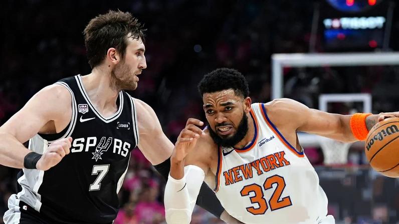Karl-Anthony Towns, wearing a white New York Knicks jersey, makes a dribble past Luke Kornet, who is in a black San Antonio Spurs jersey, during a game.